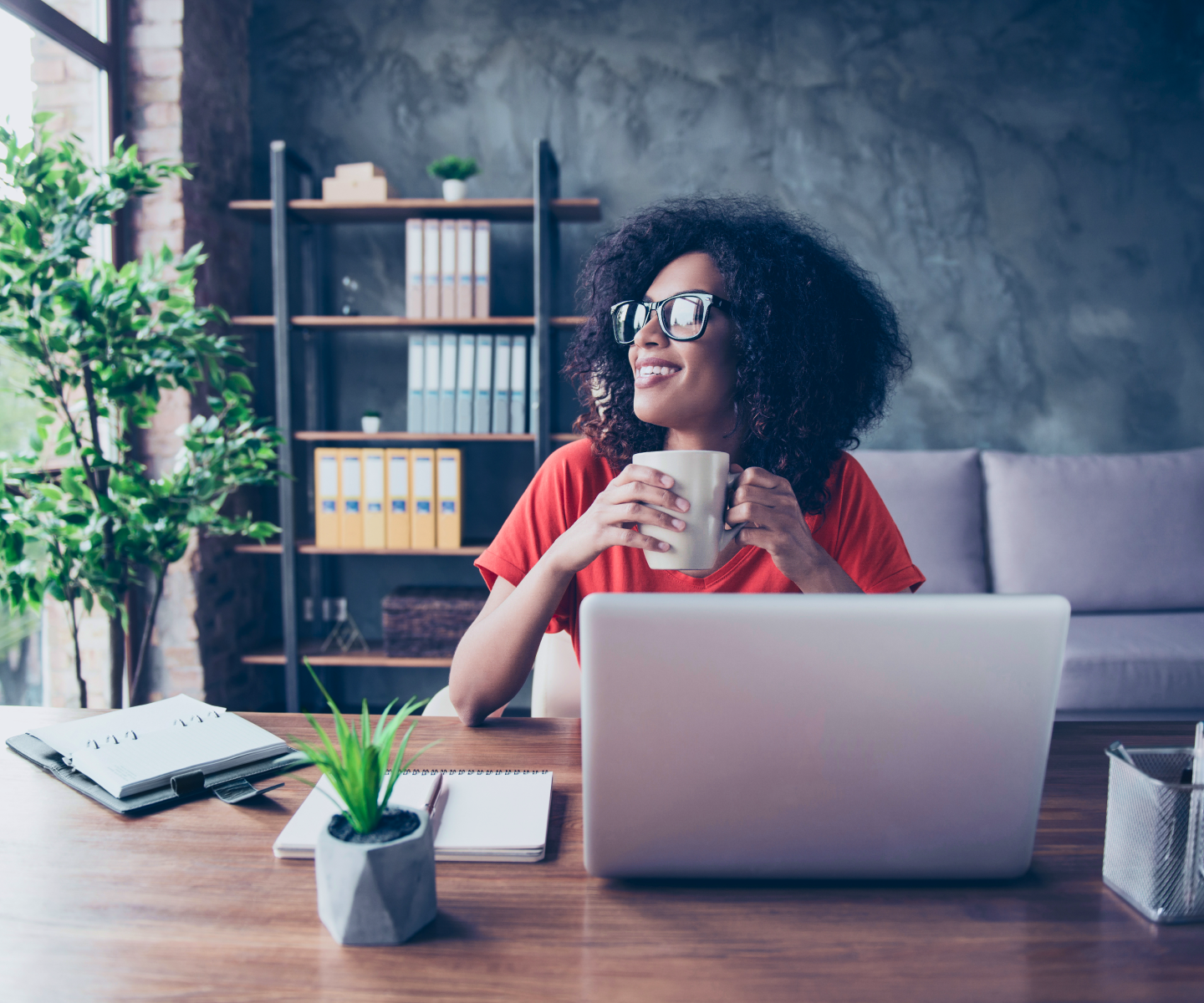 A woman with curly hair and glasses holding a coffee cup and smiling as she works at home.
