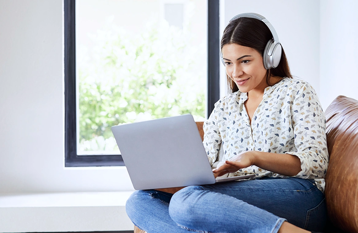A woman wearing headphones and using her laptop while sitting on the couch