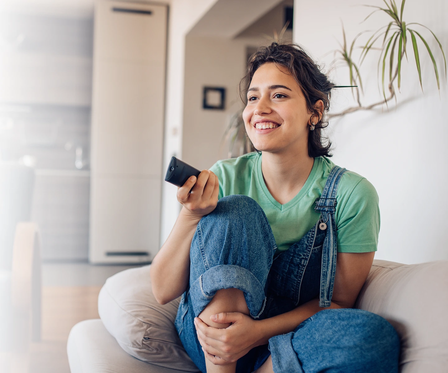 A young woman with a joyful expression sits comfortably on a light-colored sofa, holding a TV remote.